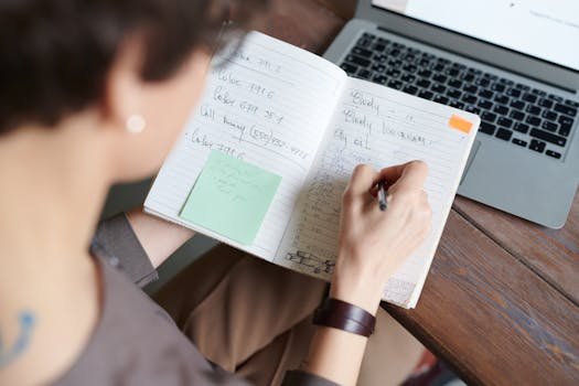 An adult writing in a notebook with a laptop on a wooden table for work or leisure.