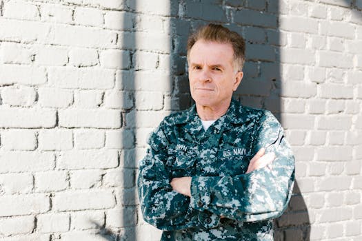 Senior Navy veteran standing proudly in naval uniform against a brick wall, outdoors in daylight.