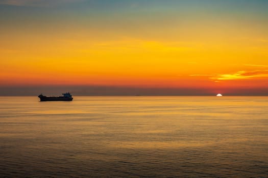 Peaceful sunset over calm ocean waters with a silhouette of a cargo ship on the horizon.