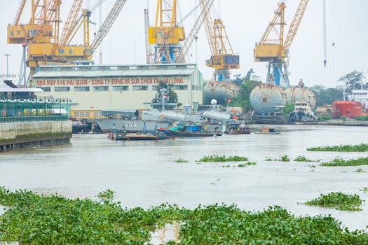 A bustling Vietnamese shipyard with cranes, military ships, and vibrant greenery.