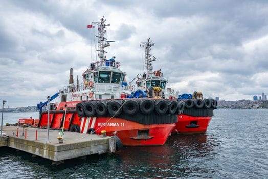 Cum să Alegi Eficient Cursul Operator GMDSS în România: Sfaturi și Costuri 2026 2 Two red tugboats docked under a cloudy sky at an urban harbor, ready for action.