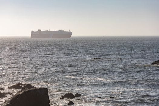 Cum să Completezi Cursul STCW Avansat Pompieri Maritimi: Cerințe și Pași Esențiali 4 A cargo ship sails on a calm ocean under a clear sky, viewed from a rocky shore.