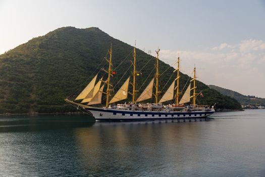 Cum să Îți Maximizezi Eficiența cu un Curs GMDSS Radiotelefonist Avansat în România 2 A majestic tall ship sails by lush green hills under a clear sky, offering a serene seascape view.