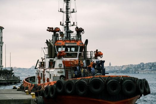 Cum să Îți Maximizezi Eficiența cu un Curs GMDSS Radiotelefonist Avansat în România 3 A tugboat moored at a harbor with a cityscape backdrop on a cloudy day.