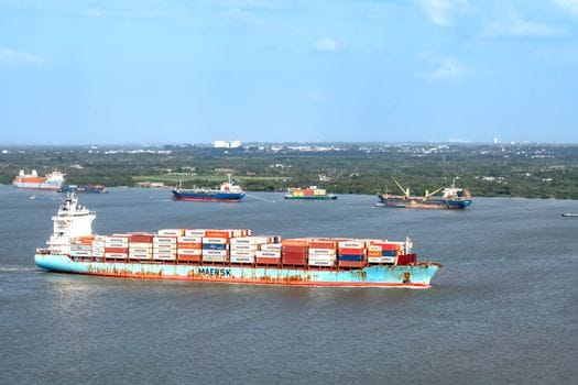 Cum să Maximizezi Oportunitățile de Angajare prin Curs Ofițer Mecanic Specializat 3 Container ships navigate a broad river with a distant cityscape under a clear blue sky.