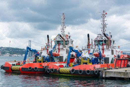Cum să Profităm de Programul STCW pentru Navigatori Peste 50 Ani 3 Vibrant tugboats moored in a bustling harbor, set against a cloudy sky and iconic bridge.
