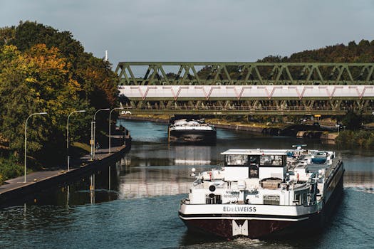 Large river barge navigating a scenic European canal beneath an iron bridge.