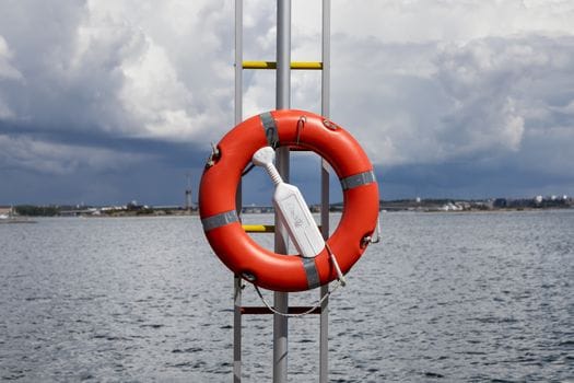 Pași pentru Pregătirea Eficientă la Examenul Radiotelefonist GMDSS: Cerințe și Beneficii pentru Navigatorii Români 2 Vibrant orange lifebuoy on a ladder by the Helsinki seashore under cloudy skies.