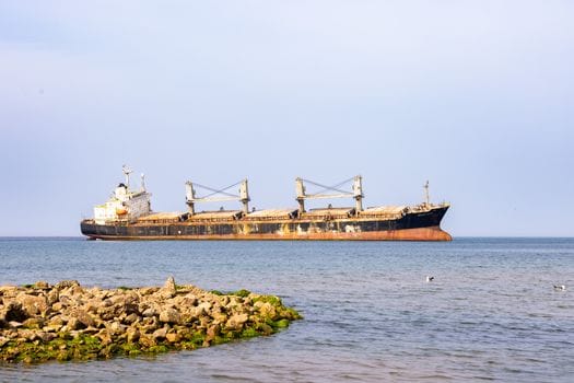 A rusting freighter docked near a rocky shoreline against a calm sea backdrop.