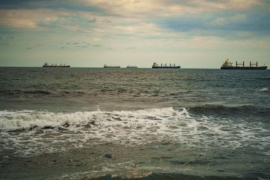 Calm ocean waves with cargo ships in the distance under a cloudy evening sky.