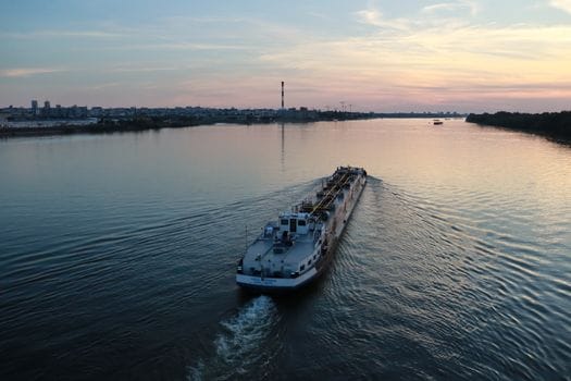Care Sunt Cele Mai Apreciate Certificări STCW de către Navigatorii Români în Iulie 2026? 2 A cargo ship travels on a calm river at sunset with a city skyline in the background.