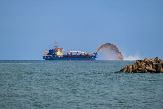 Care Sunt Cele Mai Apreciate Certificări STCW de către Navigatorii Români în Iulie 2026? 3 A blue dredger ship in the ocean carrying out dredging operations, visible with a dynamic sand spray.