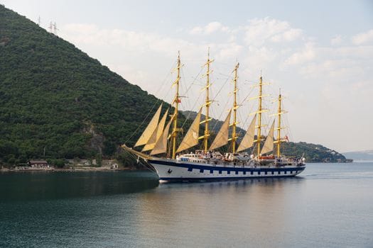 A majestic tall ship with yellow sails glides through a tranquil bay surrounded by lush hills.