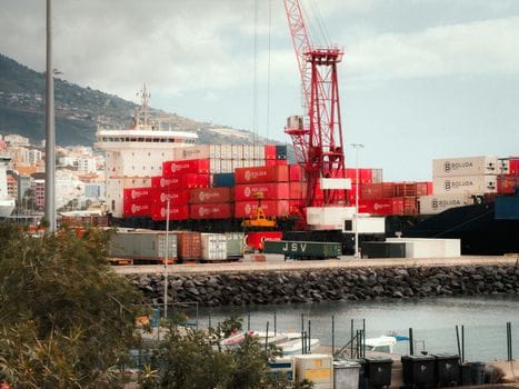 A bustling shipping port in Spain featuring vibrant red containers and a towering crane against a scenic backdrop.