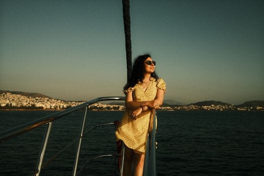 Woman in a yellow dress gazing at sea views from yacht railing, coastal backdrop.