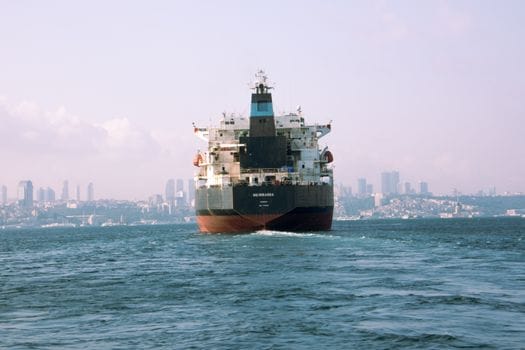 Top 8 Oportunități Educative STCW pentru Marinarul Român, Iulie 2026 4 Cargo ship sailing in open water with urban skyline in the distance.