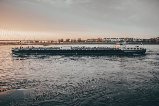 Academia Maritimă Admitere 2026 vs Certificat STCW Rapid: Ce Alegere Te Avantajează? 4 A cargo barge navigates the river under a beautiful sunset with a bridge and town in the background.