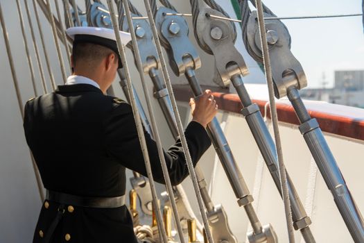 A sailor in uniform adjusts ropes on a ship deck during the day.
