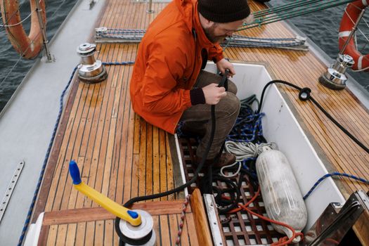 A man in an orange jacket tends to ropes and equipment on a wooden sailboat deck.