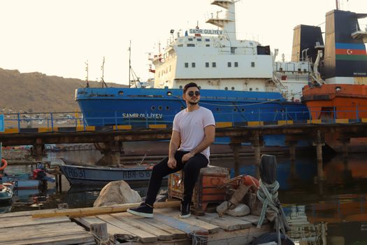 Top 5 Oportunități de Angajare ca Ofițer Secund pe Nave Comerciale în România, August 2026 2 Portrait of a young man sitting on a dock with ships and containers at a bustling port.