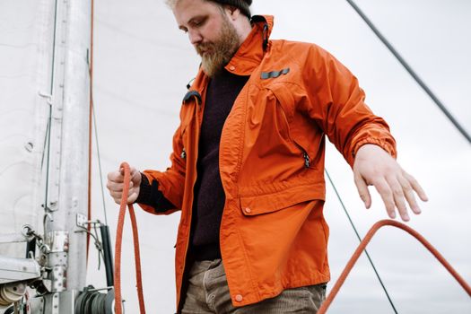 A bearded man in an orange jacket skillfully manages sailing ropes on a boat.