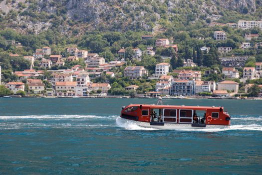 Red ferry boat cruising in front of a quaint village by the waterfront, surrounded by lush hills.