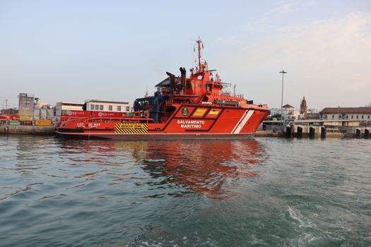 Vibrant red maritime rescue ship docked at Cádiz port against a cityscape backdrop.