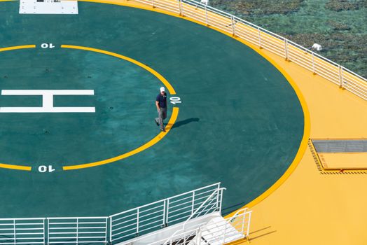 Overhead view of a man walking on a brightly colored helipad situated on a maritime or coastal platform.