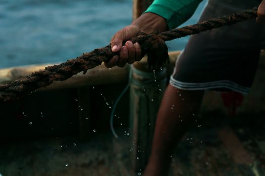 A close-up of a hand gripping a wet, textured rope on a boat, with water droplets in focus.