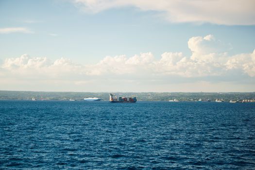 A cargo ship sails on the ocean with a scenic horizon and bright clouds in the sky.