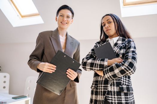 Two confident businesswomen in stylish suits holding folders in a modern office setting.