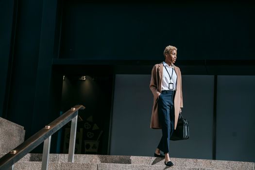 From below full body positive young female entrepreneur in stylish brown coat with hand in pocket and bag walking down stairs near office building