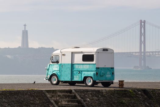 A retro van parked by the Tagus River with Lisbon's 25 de Abril Bridge in the background.
